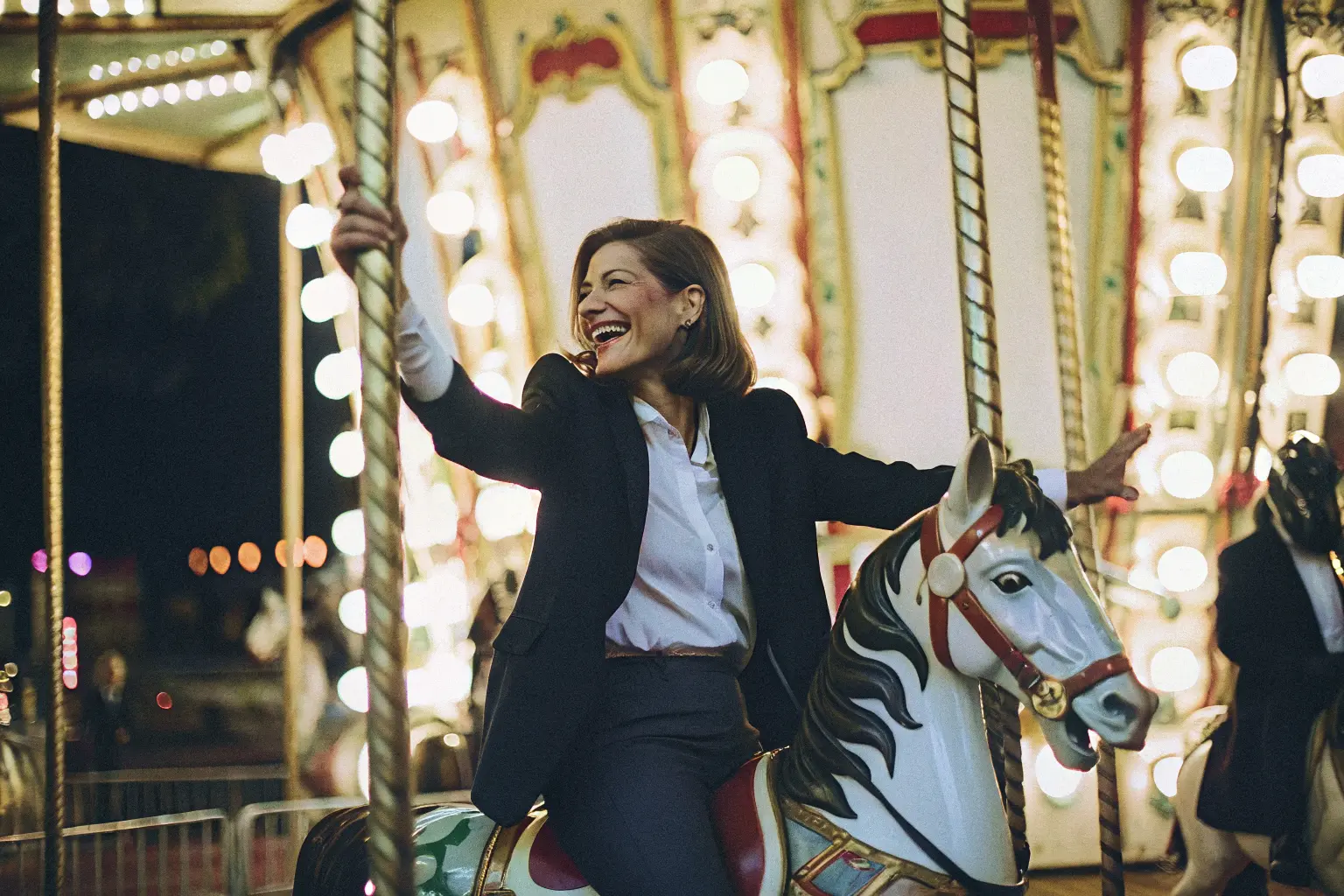 A woman wearing a suit smiles joyfully whilst riding a carousel horse at night, surrounded by bright lights and festive decorations, highlighting the importance of play in therapy for fostering happiness and wellbeing.