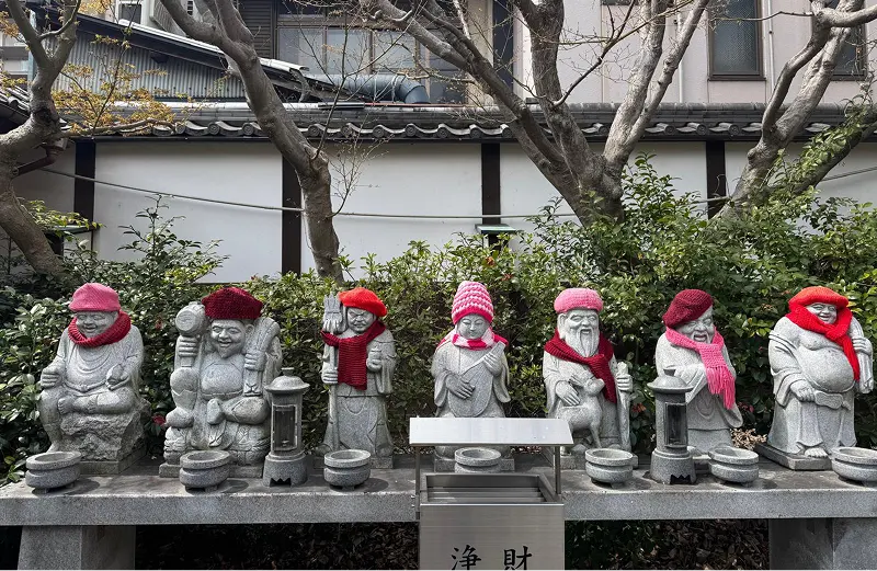 Seven stone statues with red and pink knitted hats and scarves stand in a row outdoors, in front of a low wall and trees, evoking serenity—reminiscent of the calm often sought at Therapeutic Innovation Harley Street. A donation box and lanterns sit before them.