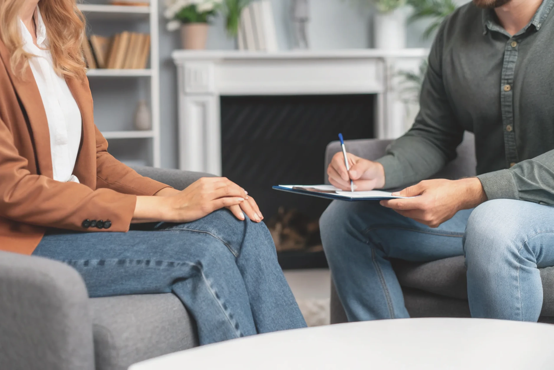 Two people sit and talk in a comfortable room, one holding a clipboard and pen while the other listens. Both are dressed casually in an inviting office or therapy space with a fireplace, reflecting the welcoming atmosphere of ADHD support London.