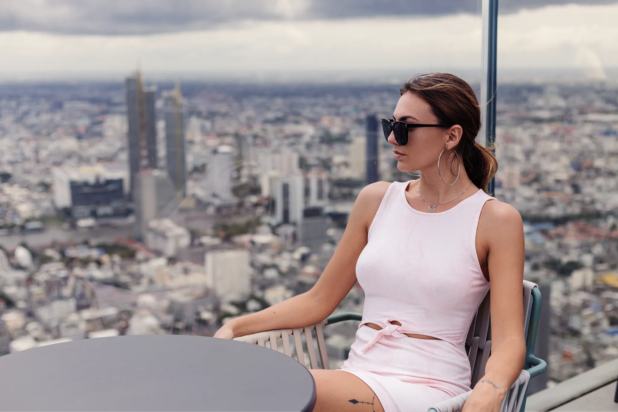 A woman in a light pink dress and sunglasses sits at a rooftop table, looking to the side, with a blurred cityscape and skyscrapers in the background under a cloudy sky—capturing the essence of Summer Anxiety Support London.