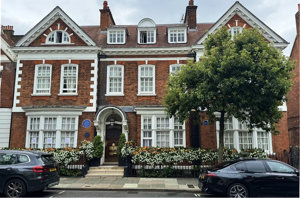 A large, red-brick townhouse with white-trimmed windows and a columned entrance, home to the Harley Street Therapy Clinic, is surrounded by blooming white flowers and greenery. Two cars are parked on the street in front of the building.