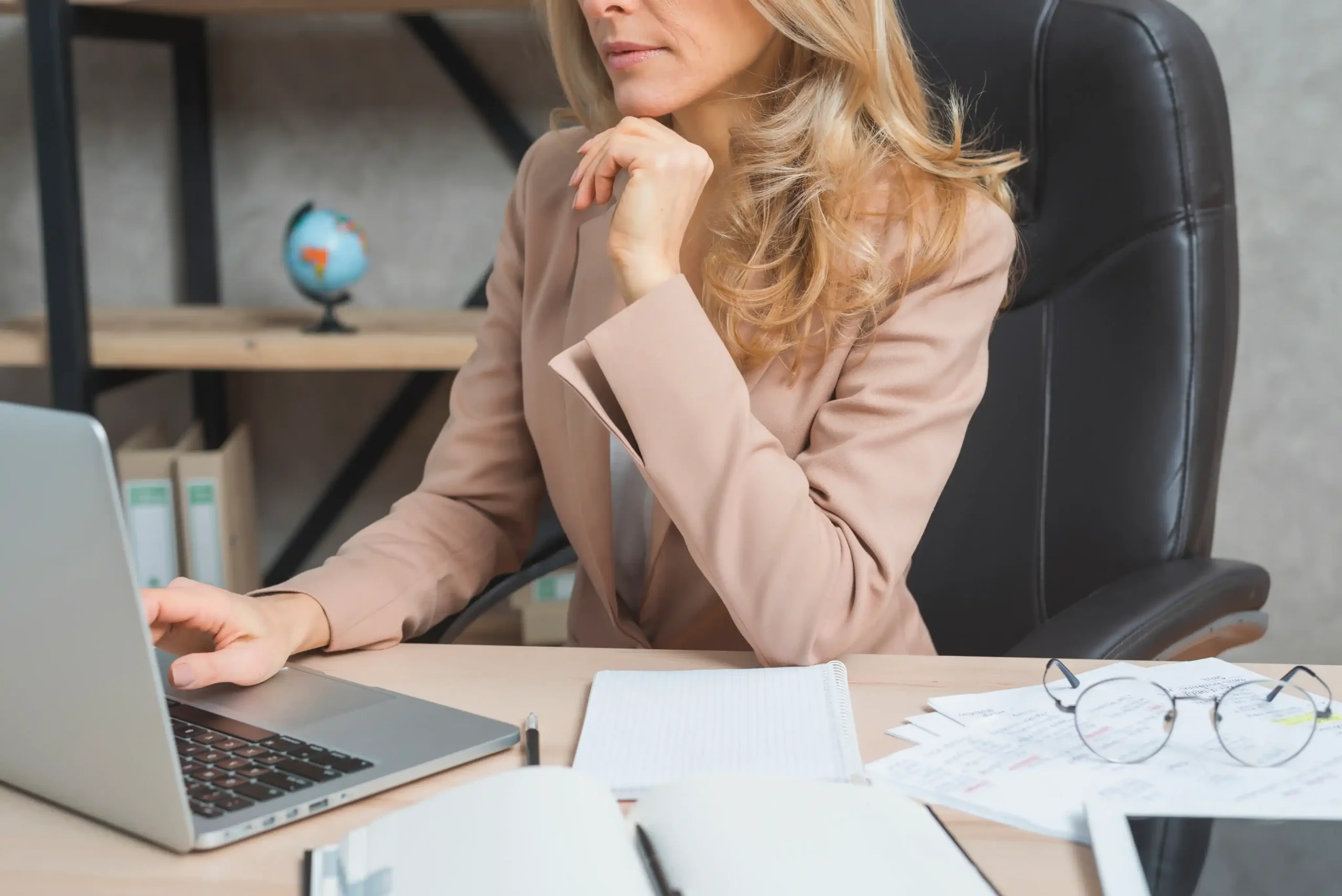 A woman in a beige blazer sits at a desk with a laptop, notebook, and papers, thoughtfully looking at her screen—possibly reflecting on workplace mental health in London. A small globe rests on a shelf in the background.