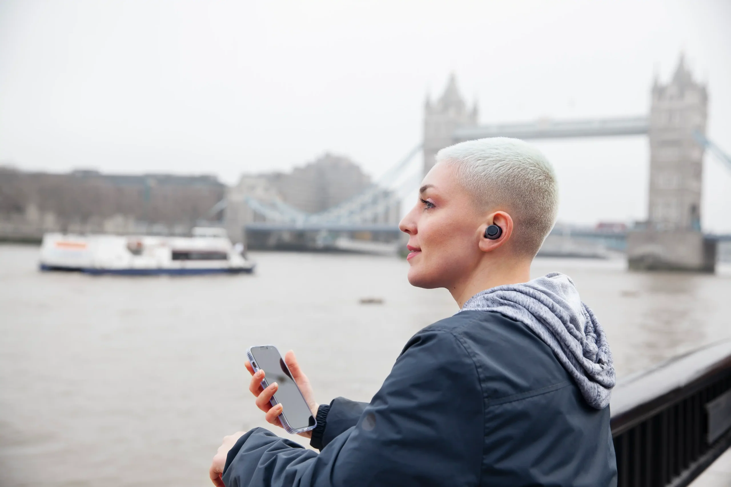A person with short, light-coloured hair wearing earphones holds a mobile phone while standing by the River Thames, building emotional resilience as Tower Bridge rises in the background on a cloudy day.