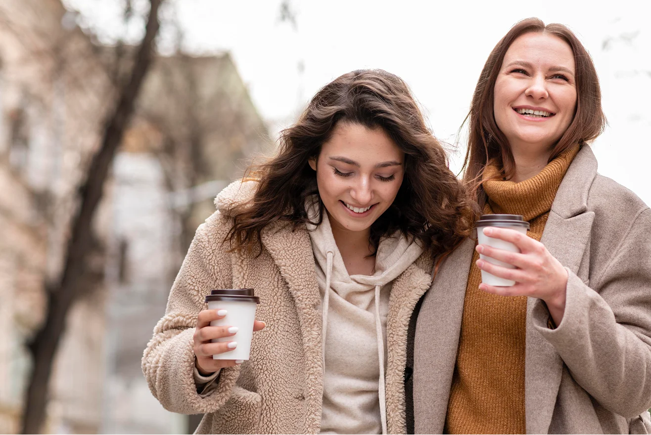 Two young women in cosy coats walk outside on a chilly day, smiling and holding takeaway coffee cups, enjoying each other's company—like friends catching up after relationship therapy London. Blurry trees and buildings are visible in the background.
