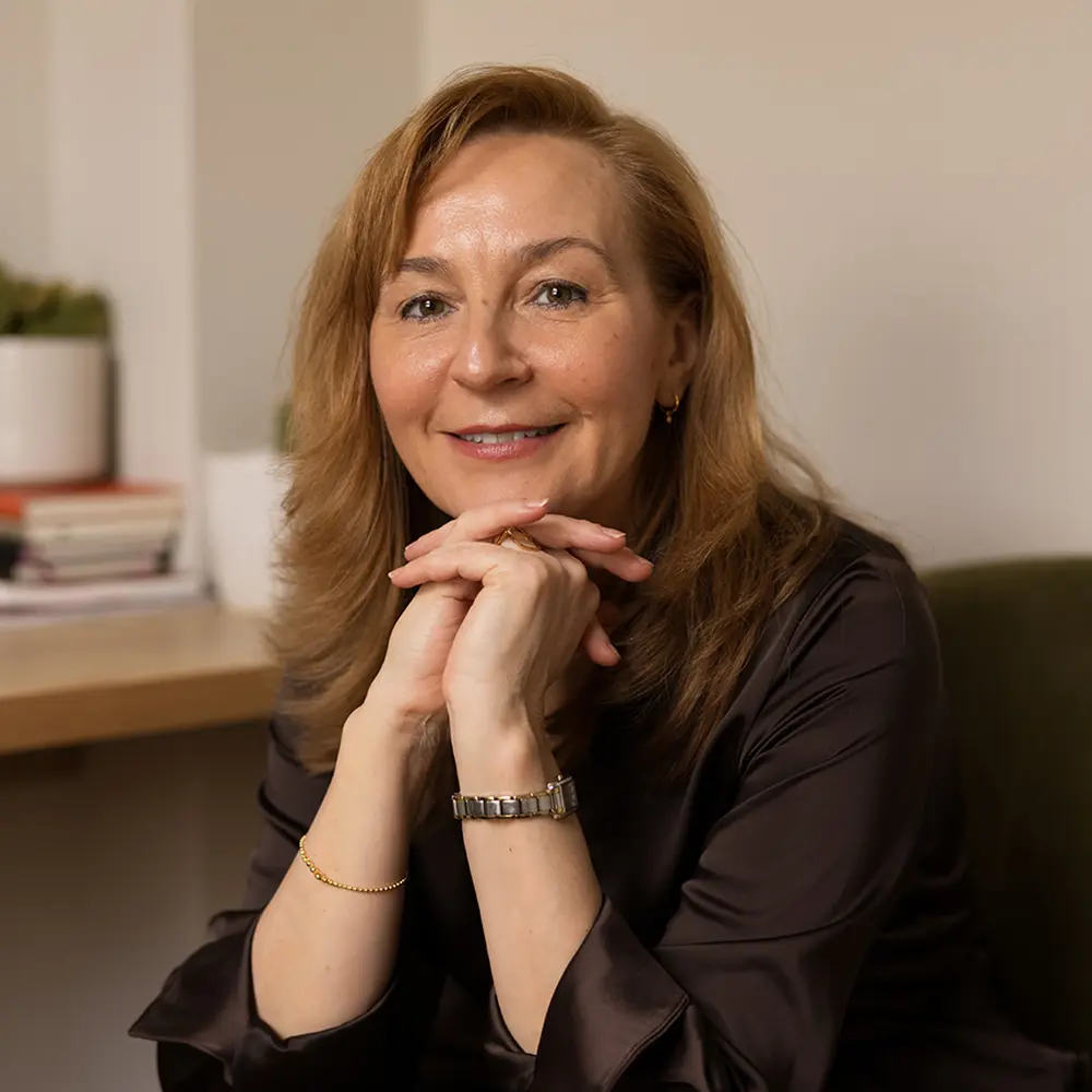 A woman with light brown hair and a dark blouse sits indoors, smiling with her hands clasped under her chin. A stack of books and a potted plant are on the desk. Olena Edwards-Skadowska, London Trusted Therapy expert, is pictured. Olena Edwards-Skadowska London Trusted Therapy Director
