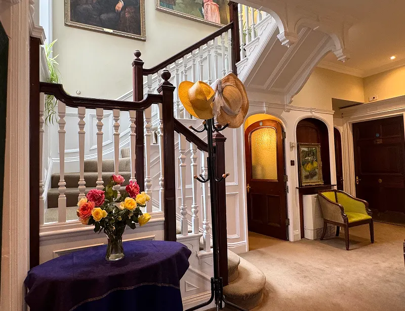 Harley Street Therapy London A cosy hallway with a staircase, a vase of colourful roses on a round table covered with a purple cloth, and a coat stand holding two hats. There’s an armchair, wooden doors, paintings on the walls—reminiscent of Harley Street’s inviting calm.