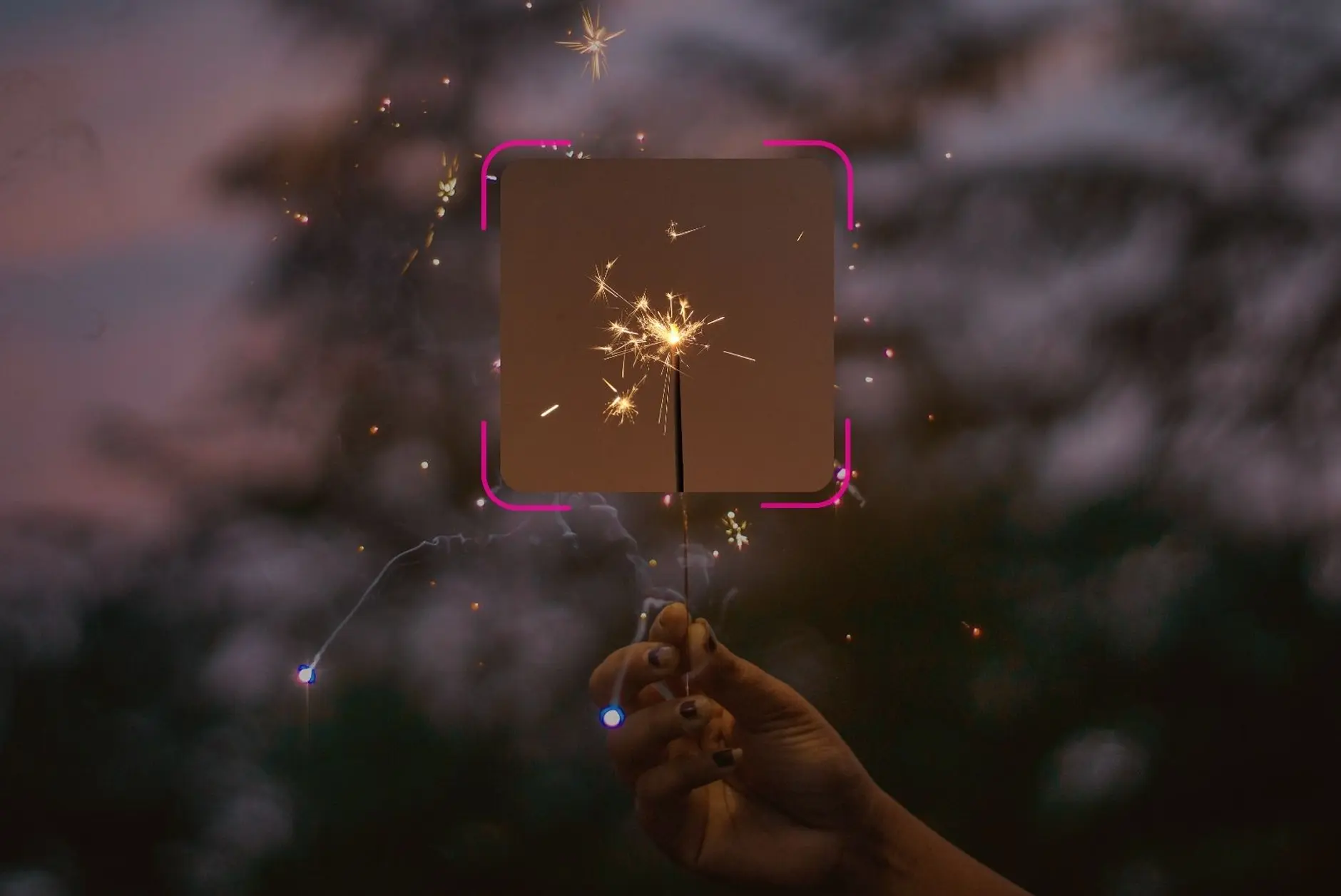 A hand holding a lit sparkler at dusk, sparks flying—a blurry background of dark trees sets the mood. A pink, square-shaped digital overlay highlights the sparkler, capturing a fleeting moment of Christmas anxiety and celebration.