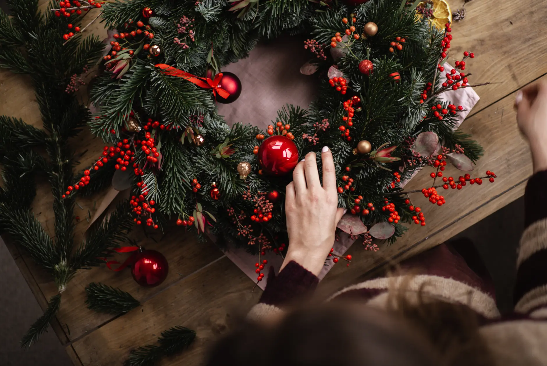A person decorates a festive Christmas wreath with red berries, baubles, and pine branches on a wooden table. Viewed from above, their hand adjusts the wreath with care, hoping to ease any Christmas performance anxiety about holiday decorating.