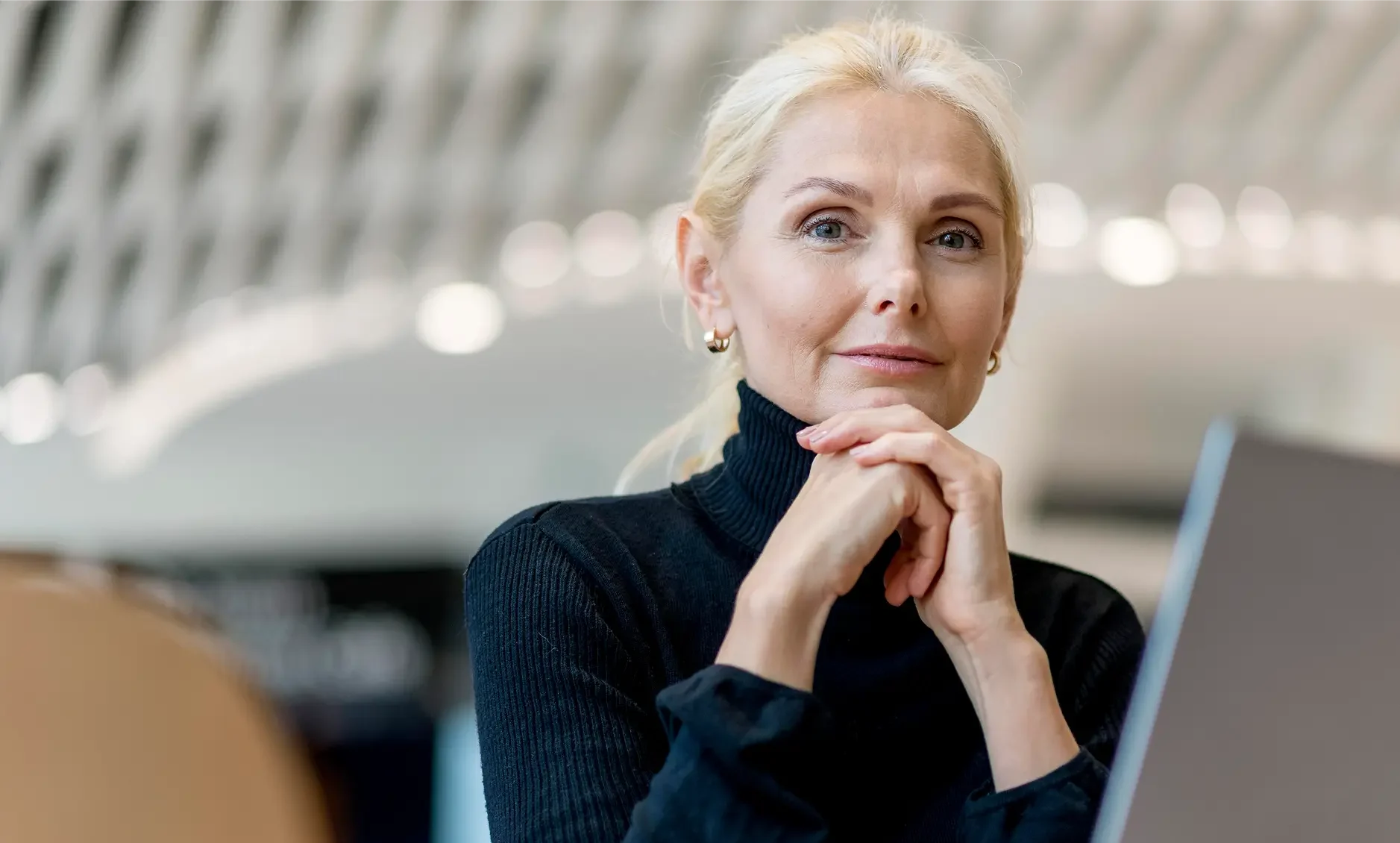 A woman with blonde hair in a ponytail, wearing a black polo neck, sits indoors with her hands clasped under her chin, looking thoughtfully at the camera—perhaps reflecting on ADHD symptoms in women. A blurred background and part of a laptop are visible.