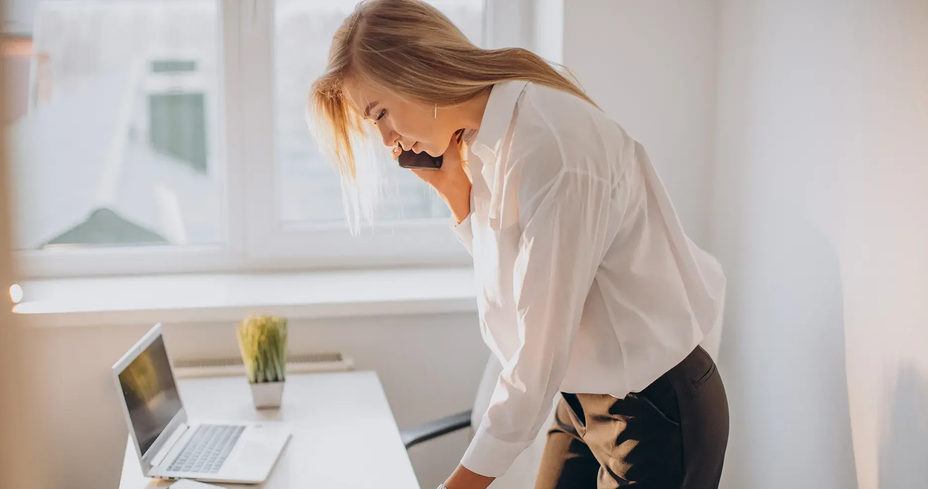 A woman in business attire stands at a desk, talking on the phone while looking at documents—navigating ADHD at Work London amid folders, a laptop, and a potted plant as sunlight streams through the window.