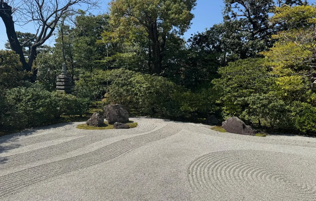 A traditional Japanese Zen garden with raked gravel patterns, large rocks, and lush green trees under a clear blue sky—a serene retreat that inspires wellbeing and healing.