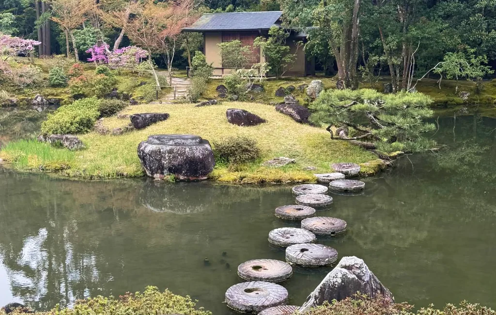 A tranquil Japanese garden with a pond, circular stone steps crossing the water, green grass, rocks, trees, blooming flowers, and a small wooden pavilion in the background offers healing beauty just outside your door.