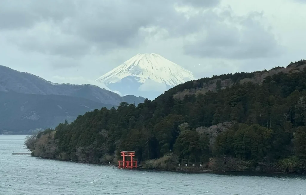 A snow-capped Mount Fuji rises in the background behind forested hills and a lake, with a bright red torii gate at the water’s edge — an inspiring scene that evokes healing and wellbeing amid cloudy skies.