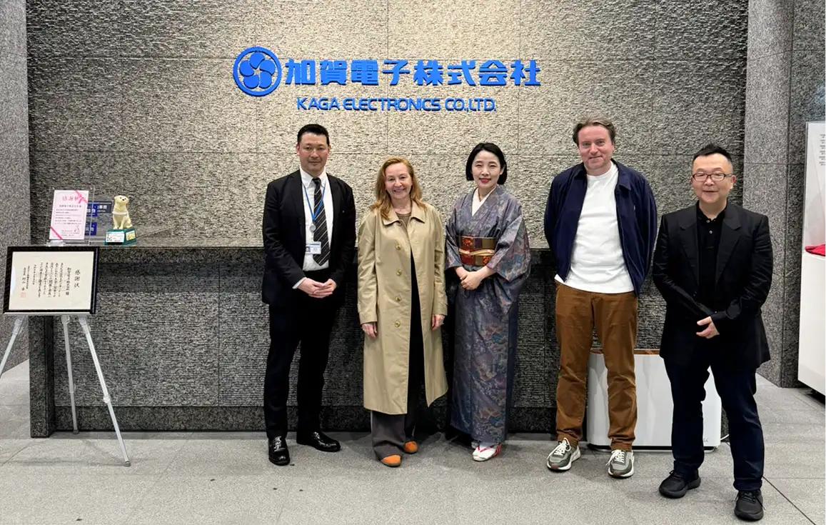Five people stand in front of a grey wall with the Kaga Electronics Co., Ltd. logo. They are all smiling, highlighting their commitment to wellbeing technology reminiscent of London Trusted Therapy and Harley Street’s renowned care standards.
