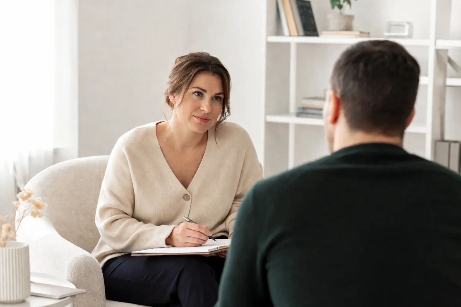 A woman sits in a chair holding a notebook and pen, attentively listening to a man opposite her in a cosy, well-lit room filled with books—an inviting space for EMDR or CBT therapy sessions focused on trauma and PTSD.