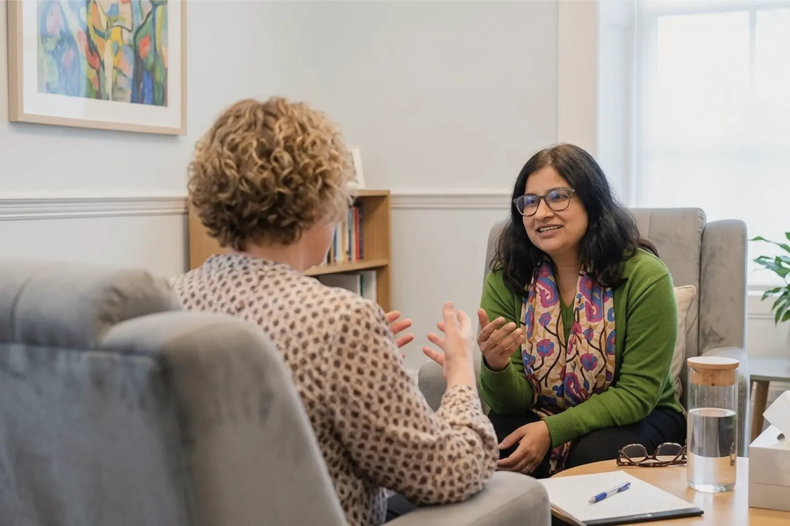 autistic-burnout-recognising-the-signs-before-crisis-point Two women sit facing each other in a cosy office; one speaks while gesturing, and the other—possibly a couples therapist London—listens and smiles, holding a pen. A notebook, glass of water, and bookcase are visible in the background.