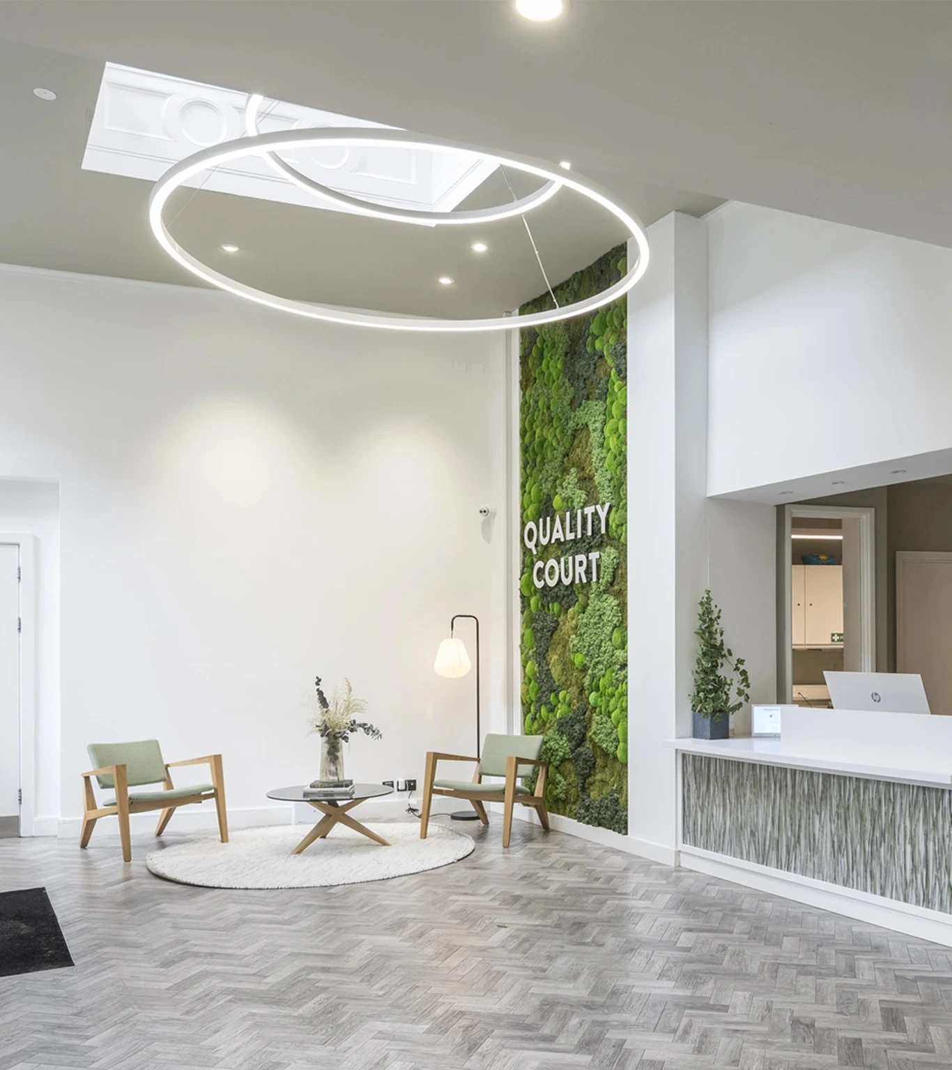 A modern lobby with light wood flooring, two green chairs around a round rug, a glass coffee table, a large moss wall with QUALITY COURT text, and a reception desk with a plant. Circular ceiling lights hang above.