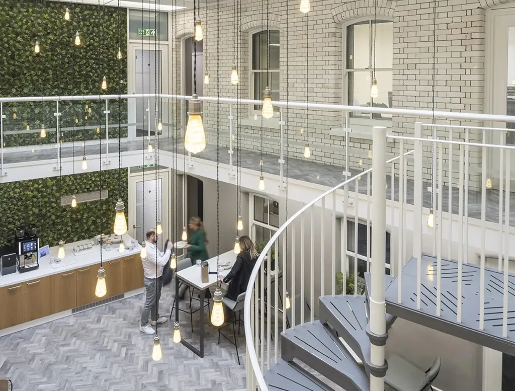 Modern office space with a spiral staircase, hanging pendant lights, a plant-covered wall, and people sitting at a table near a coffee station. The setting is bright, with white brick walls and large windows.
