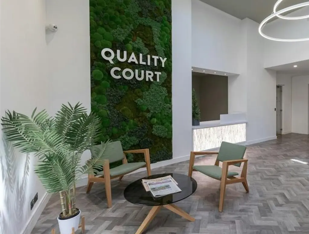 Modern lobby with three wooden armchairs around a glass coffee table, a potted plant, and a large green wall featuring the words QUALITY COURT in white letters. Reception desk and minimalist décor in the background.