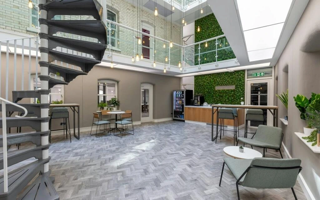 Modern, bright lounge with a spiral staircase, seating areas, vending machines, plants, and a glass ceiling. Hanging lights and large windows add natural light to the open, inviting space.