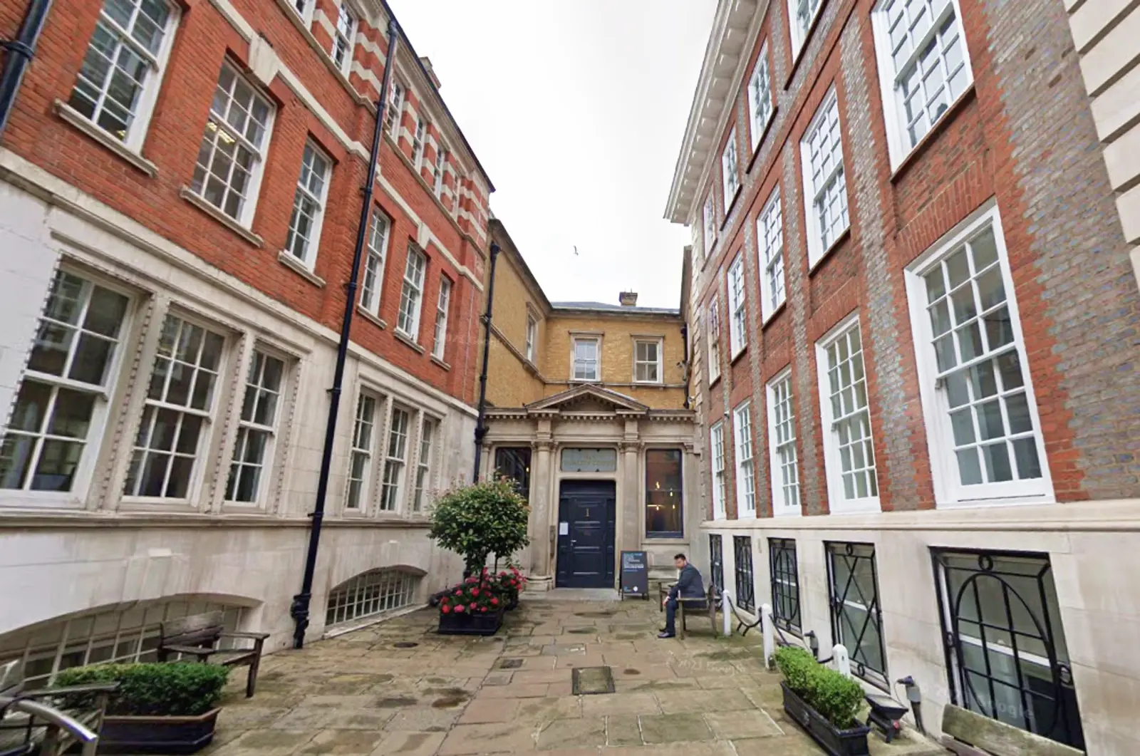 Chancery Lane Therapy London | Legal District | London Trusted Therapy A small stone courtyard with potted plants is surrounded by tall, red-brick and white-trimmed buildings with large windows; a person sits on a bench near the entrance of a building with columns.