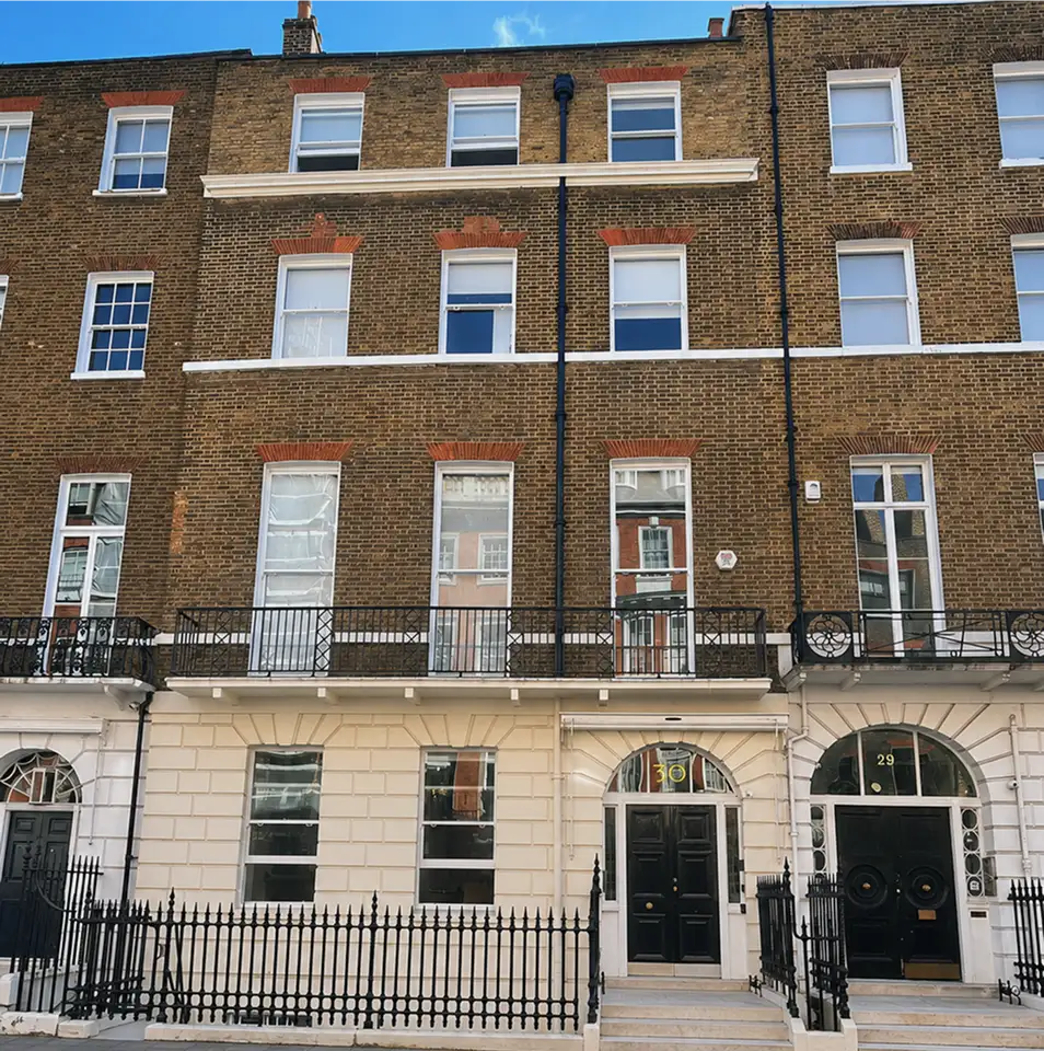 A row of classic Georgian terraced houses with brown brick upper storeys, white-painted ground floors, arched doorways, tall sash windows, black railings, and a clear blue sky above.