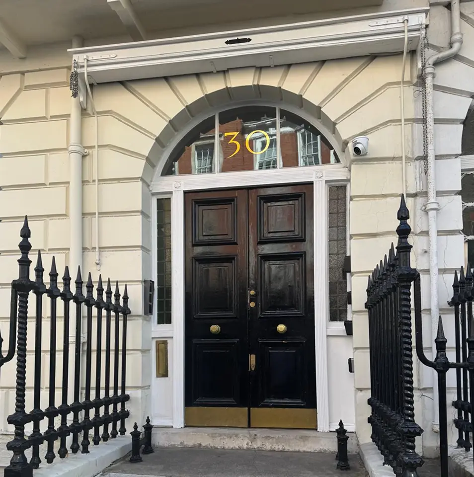 A black double door with gold accents and the number 30 above it, set in a cream-coloured building with ornate iron railings on both sides.