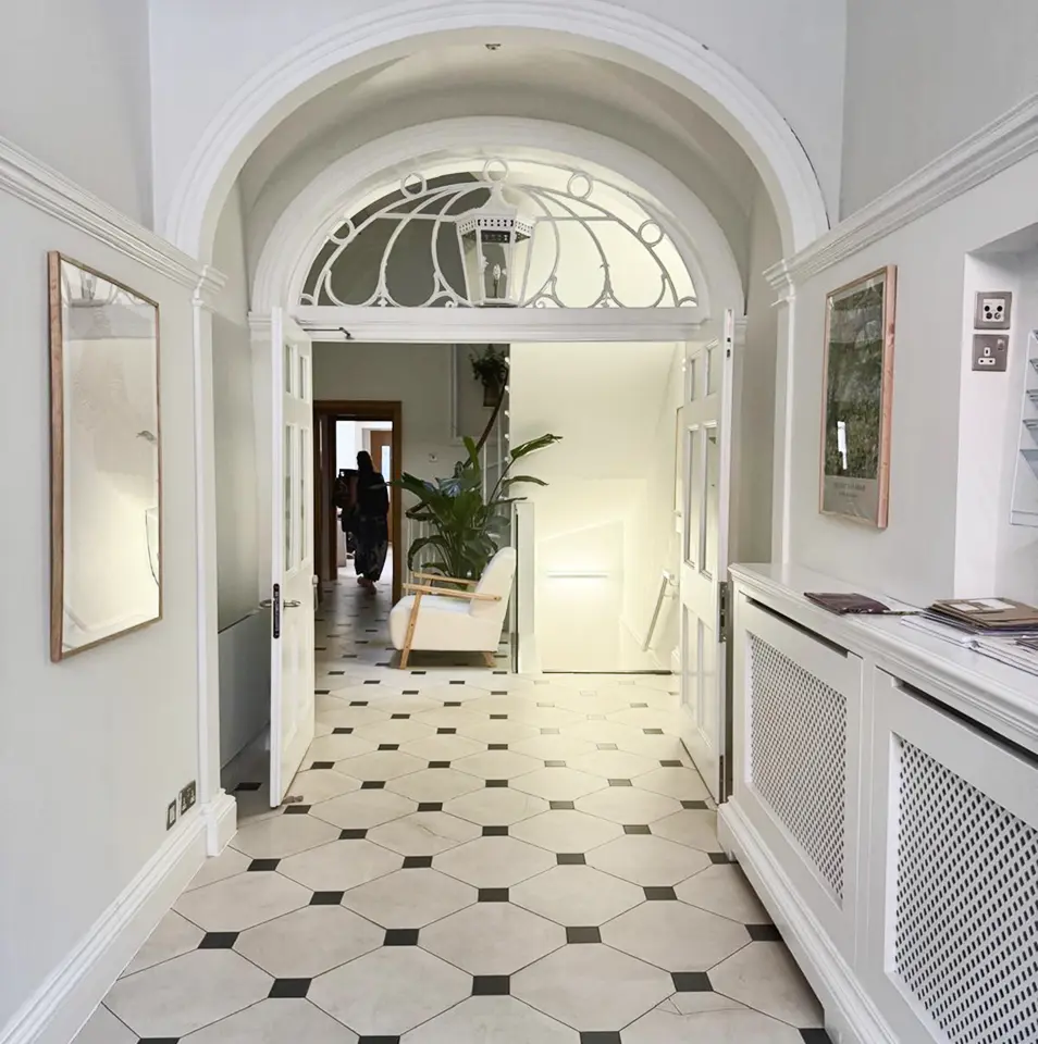 A bright hallway with white walls, black-and-white hexagonal tile flooring, arched ceiling details, and a large plant at the far end. Open double doors lead to another room, and framed pictures hang on the walls.