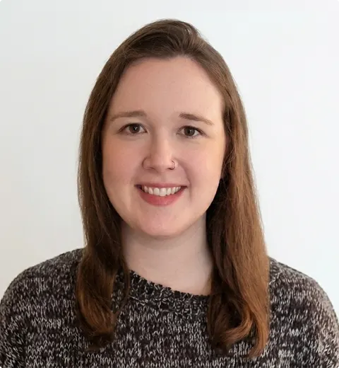 Dr Emma Corcoran | Clinical Psychologist and Therapist London A woman with straight brown hair and fair skin is smiling at the camera. She is wearing a brown and white patterned jumper and is posed in front of a plain white background.