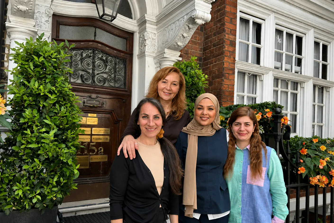 Four women stand smiling in front of a building with a wooden door and decorative plants. One woman, who appears to be a specialist SLT, stands behind the others, resting her hand on another’s shoulder. The group, possibly part of a multi-disciplinary team supporting AAC users, appears happy and relaxed.
