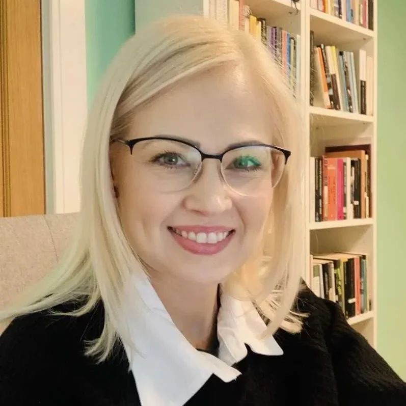 Dr Jo Parkinson Dr Jo Parkinson, a woman with blonde hair and glasses, smiles at the camera as she sits in front of a bookcase filled with books. She is wearing a black jumper with a white collared shirt underneath.